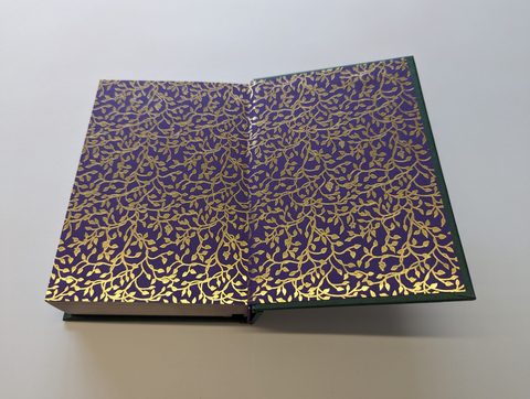 A hardback case opened flat: all-over gold foil foliage blocked on purple and green cloth
